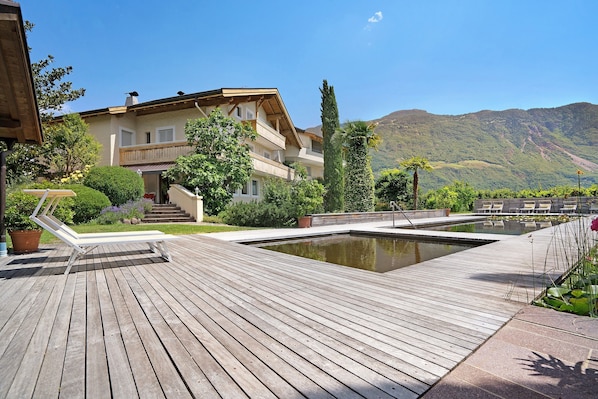 A wooden deck with a pool, a multi-story building with balconies, and a mountainous backdrop.