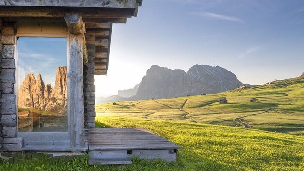 Une construction rustique en bois, dotée d’une grande fenêtre, située dans un champ verdoyant avec une montagne en arrière-plan.