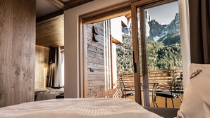 Een slaapkamer met een houten plafond, een bed met een witte dekbedovertrek, een raam met uitzicht op de bergen en een balkon met een stoel.