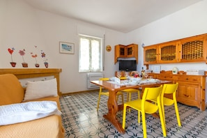 A cozy room with a bed, a dining table with yellow chairs, a kitchen area with wooden cabinets, and a patterned floor. - Apartment San Giovanni Lido Close to the Beach with Sea View & Balcony (Alghero)