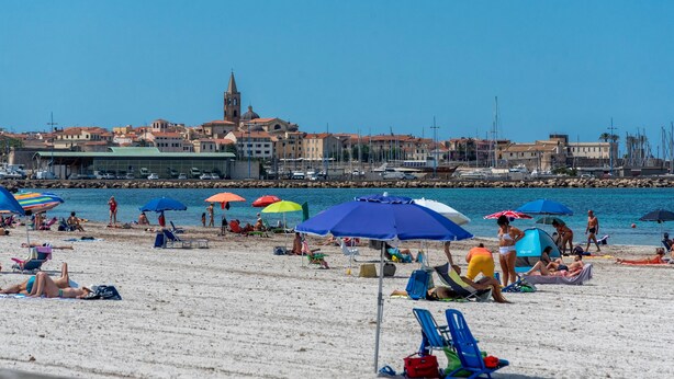 A beach with umbrellas, people sunbathing, and a building with a tower in the background. - Apartment San Giovanni Lido Close to the Beach with Sea View & Balcony (Alghero)