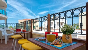 A balcony with a wooden table, yellow cushioned chairs, a yellow mug, a red cup, a basket with flowers, and a view of buildings and the sea. - Apartment San Giovanni Lido Close to the Beach with Sea View & Balcony (Alghero)