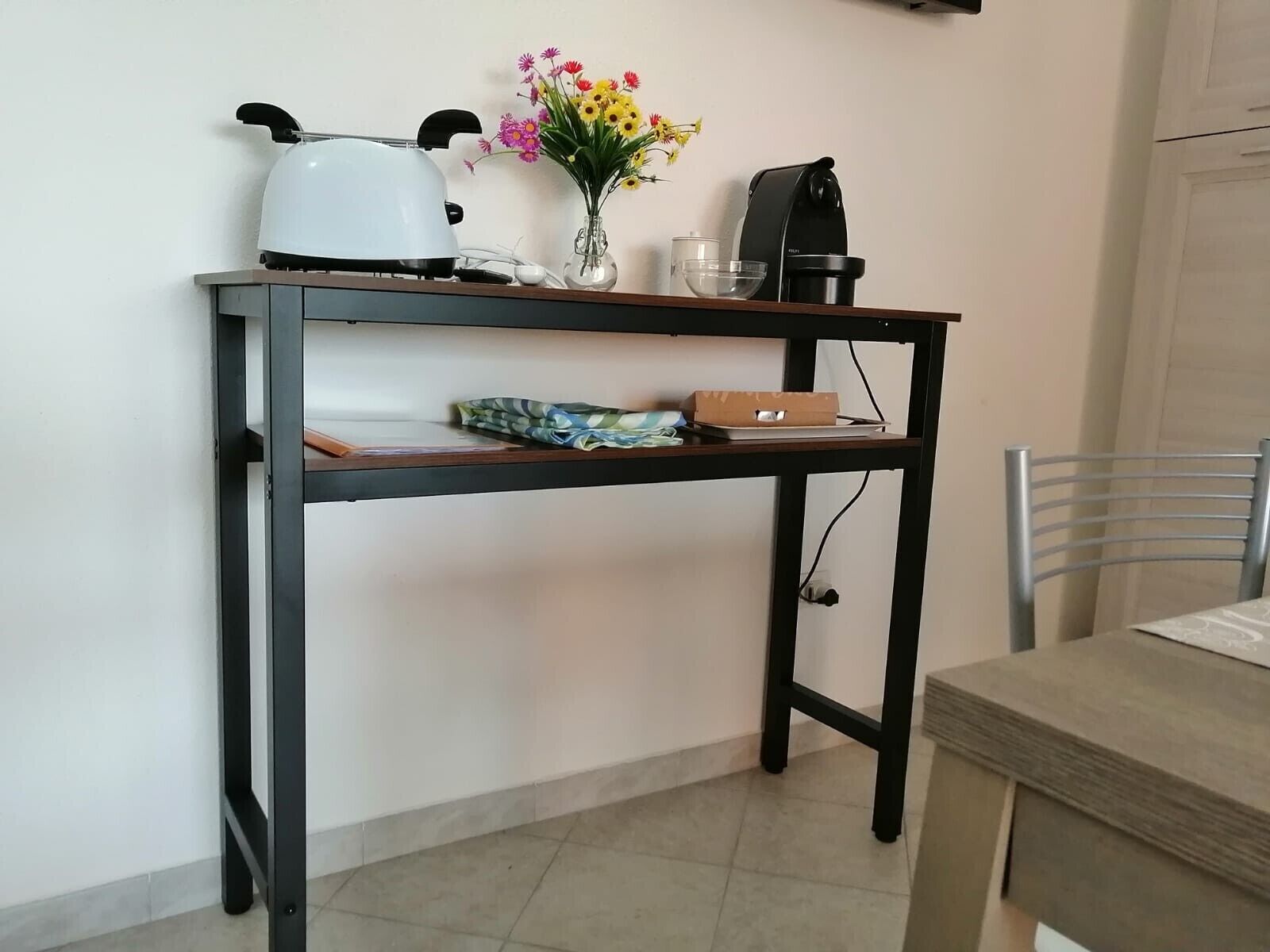 A kitchen area with a coffee maker, kettle, and a vase of flowers on a shelf.
