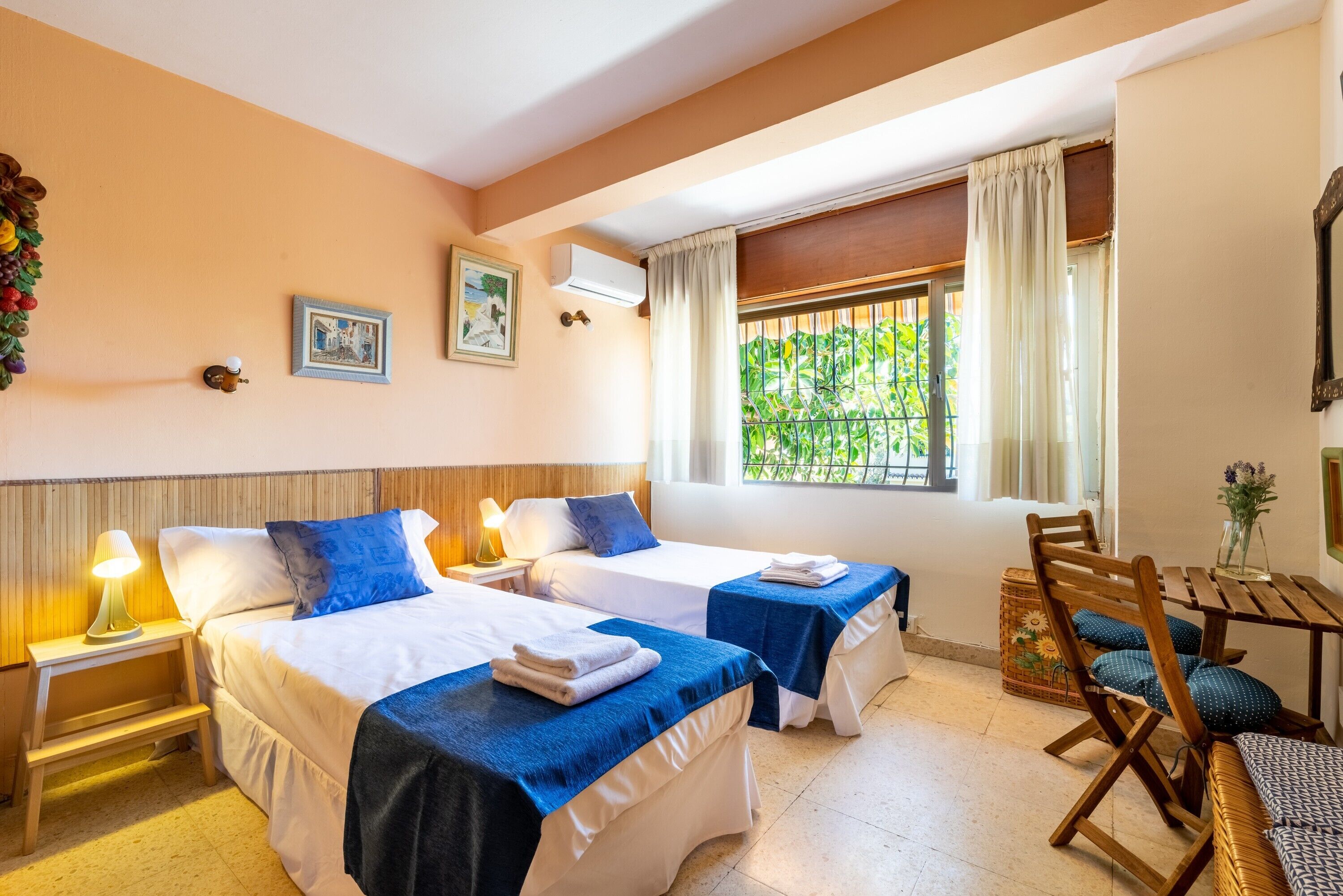 A hotel room with two beds, a small table, a chair, and a window with curtains.