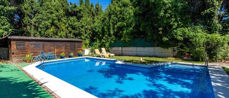 Piscina rectangular con un muelle de madera y sillas de descanso, rodeada de abundante vegetación y árboles altos.