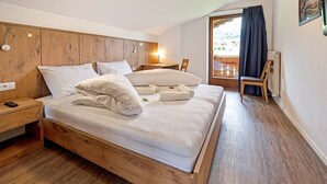 Een houten bed met witte beddengoed, een nachtkastje met een afstandsbediening, een stoel en uitzicht op de omgeving door een raam.