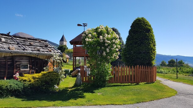 Un edificio rustico in legno con tetto spiovente, una piccola torre e un giardino recintato con un albero e una staccionata.