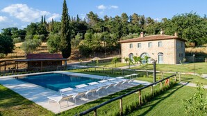 Une piscine avec des chaises longues, un bâtiment en pierre et une zone herbeuse avec des arbres.