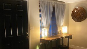 En gang med en sort dør, en træbænk med en lampe, en dekorativ genstand på væggen, og et vindue med blå persienner og hvide gardiner.