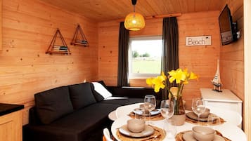 Interior acolhedor de uma cabana, com um sofá preto, uma mesa de jantar posta para quatro pessoas e uma janela com cortinas.