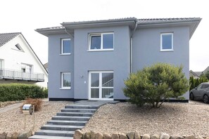 A two-story house with a grey exterior, a front door with glass panels, and a small tree in the front yard.