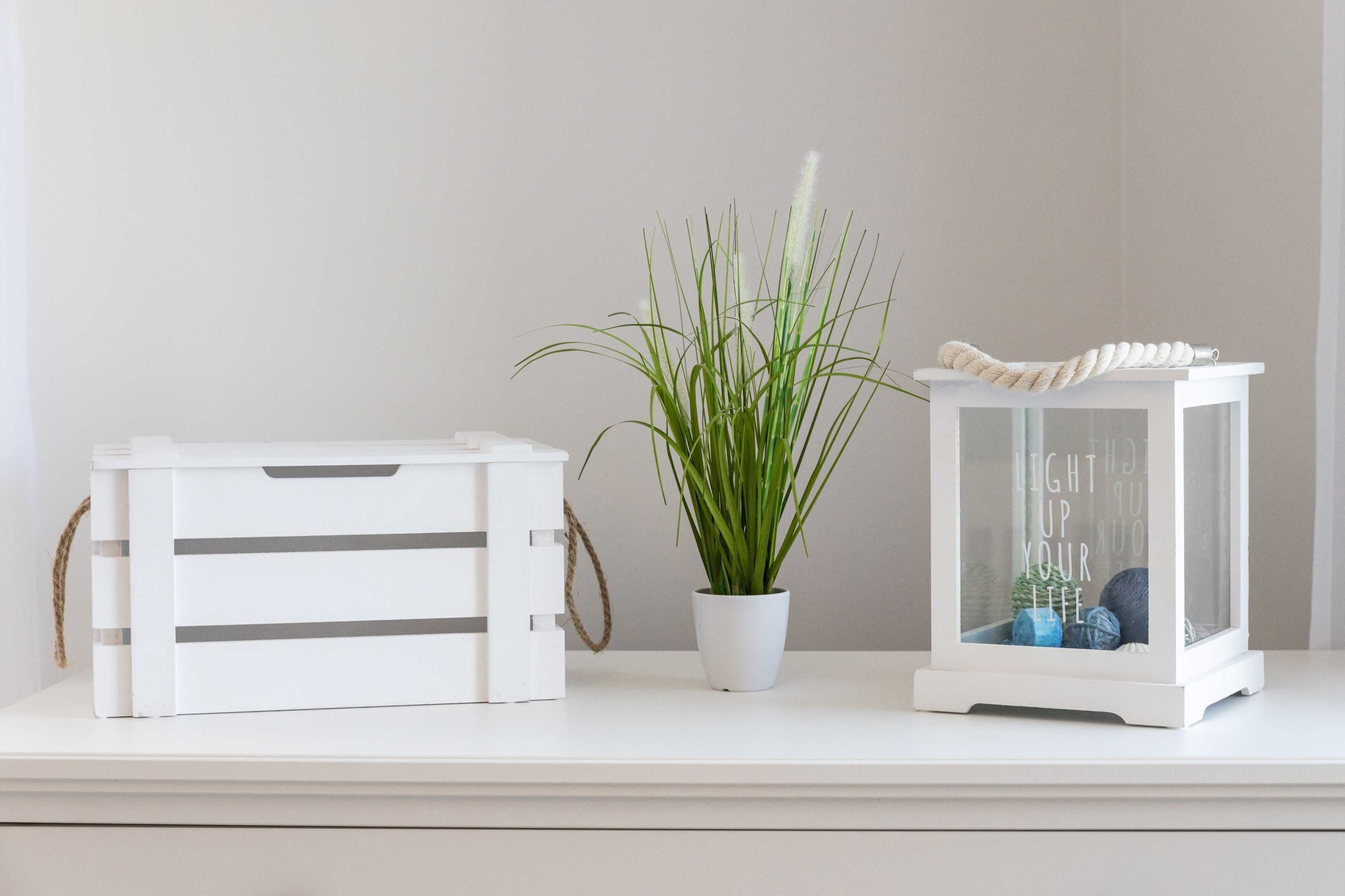 A white storage box, a potted plant, and a glass lantern on a white shelf.