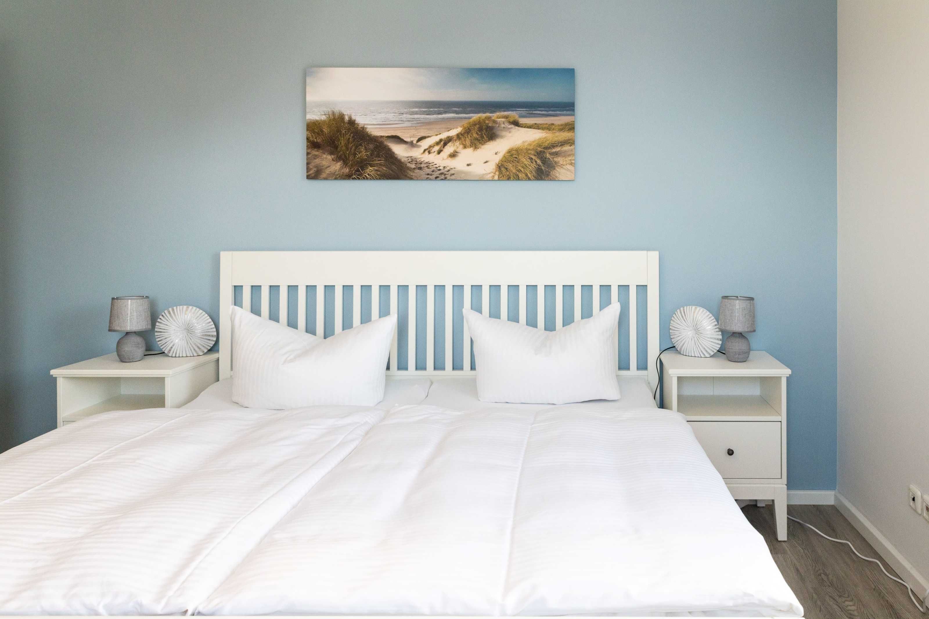 A bedroom with a white bed, two nightstands with lamps, a blue wall, and a beach scene painting.