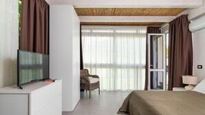 Quarto de hotel moderno com televisão de ecrã plano sobre uma cómoda branca, cama com colcha verde, cadeira de vime e uma janela grande com cortinas leves.