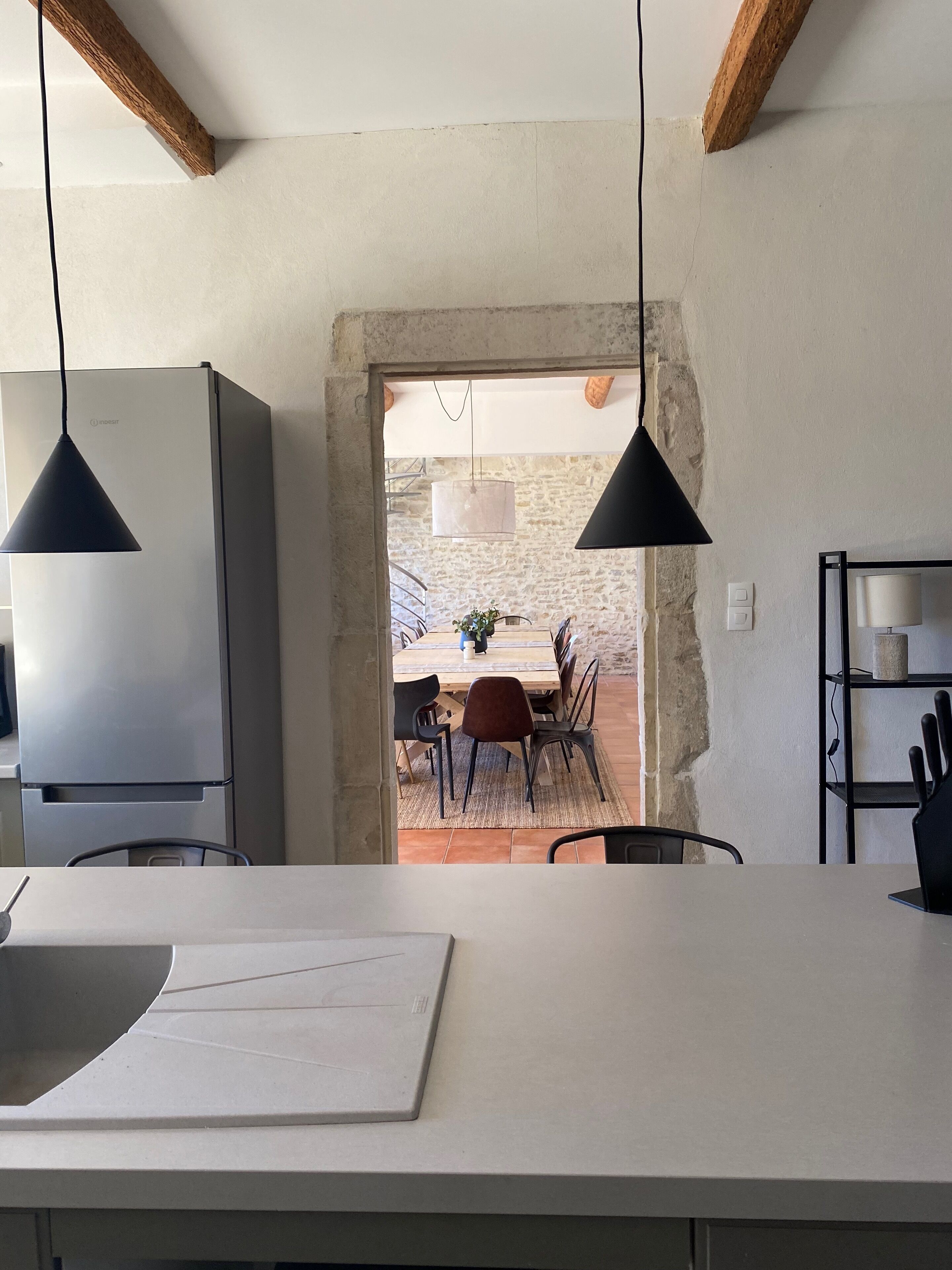 A modern kitchen with a large island, pendant lights, and a view into a dining area with a rustic stone wall.