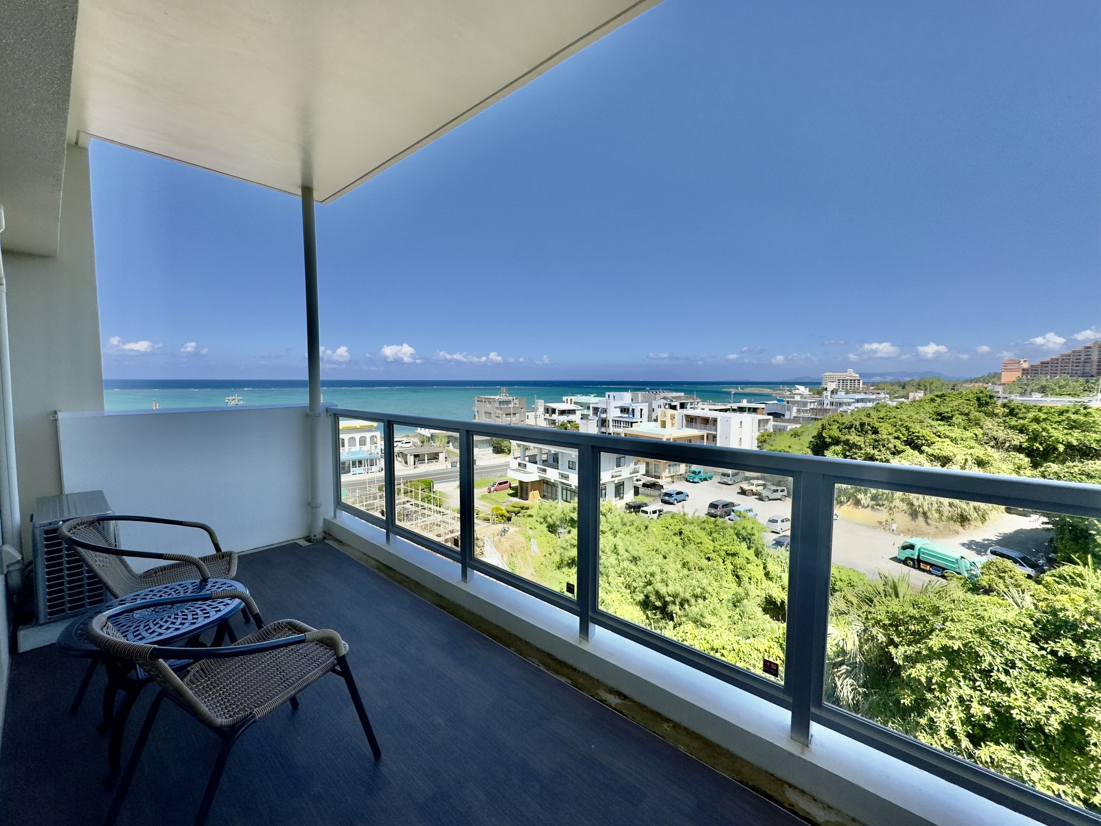 A balcony with wicker chairs, a view of the sea, and a parking area with vehicles.