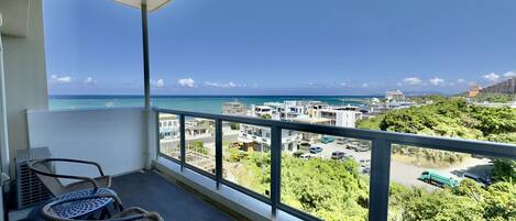 A balcony with wicker chairs, a view of the sea, and a parking area with vehicles.