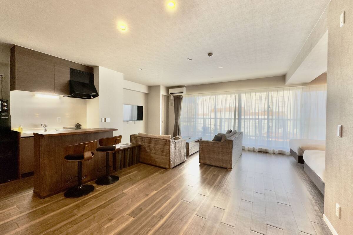Une chambre d’hôtel moderne dotée d’un coin cuisine, d’un espace salon avec un canapé et des fauteuils, et de grandes fenêtres avec des rideaux.