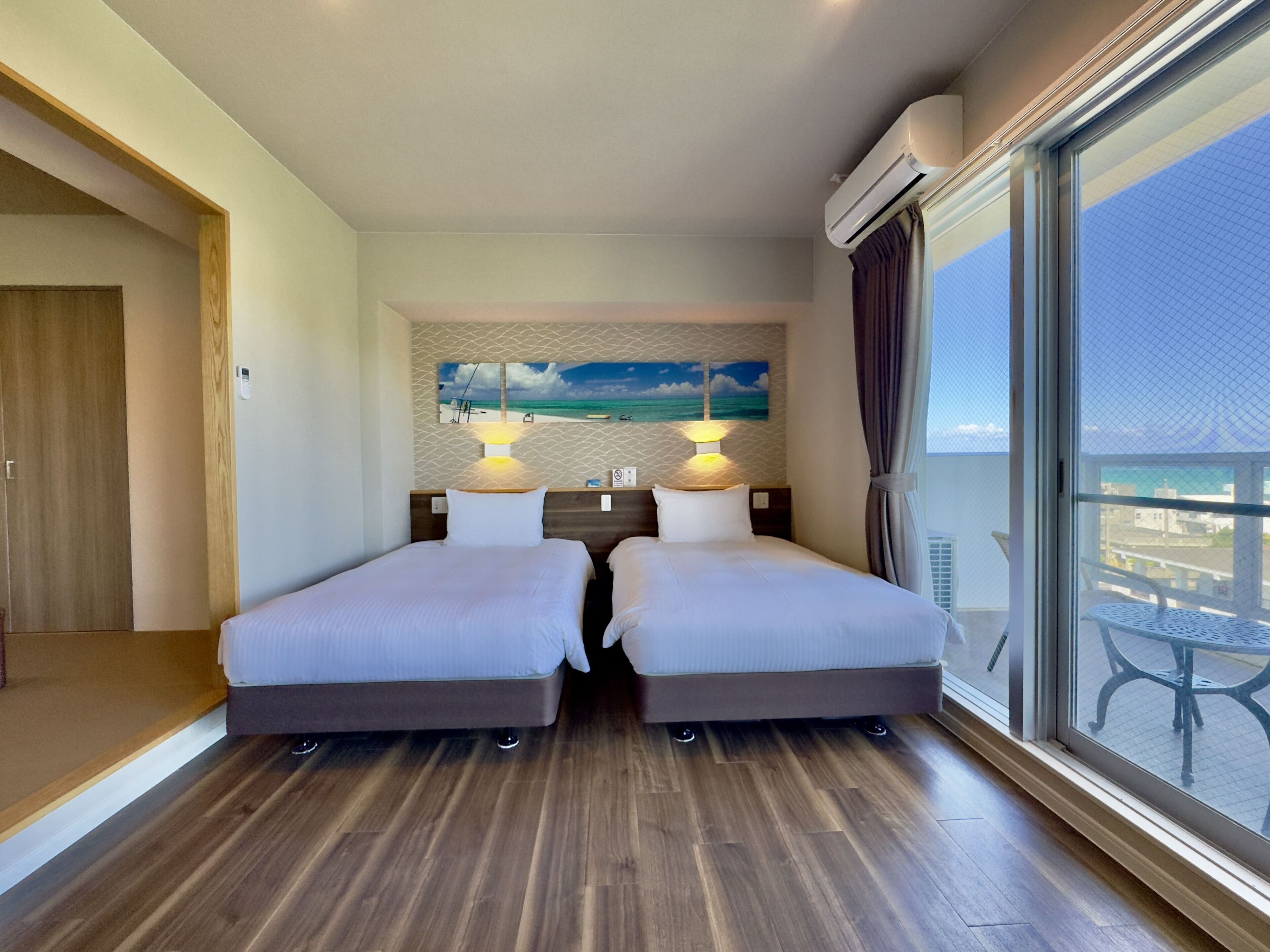 A hotel room with two beds, a large window with a view, a painting on the wall, and an air conditioning unit.