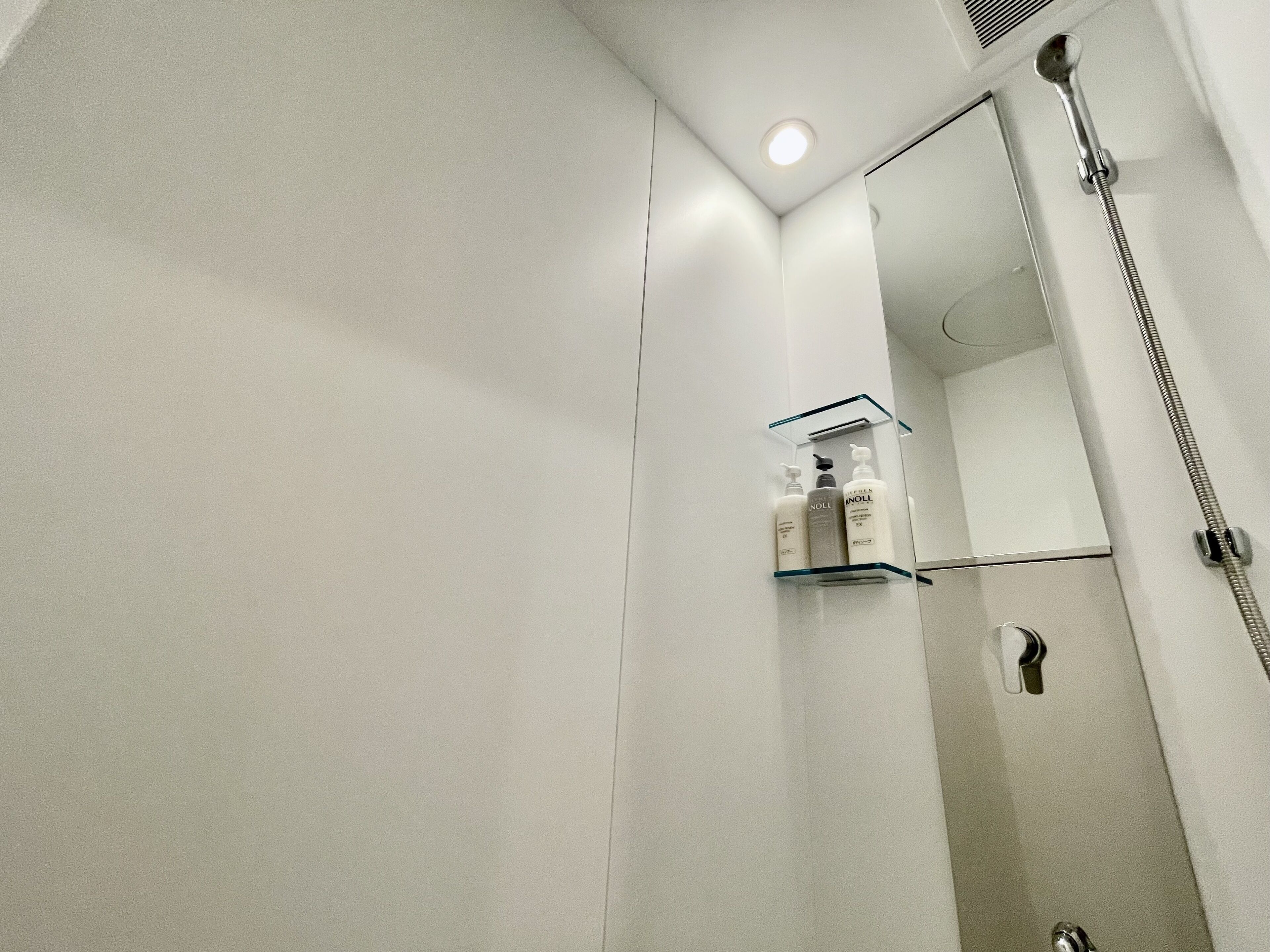 Egy modern fürőszoba, melyben üvegpolcon helyezkednek el a piperekészletek, van egy kézi zuhanyfej és egy tükör.