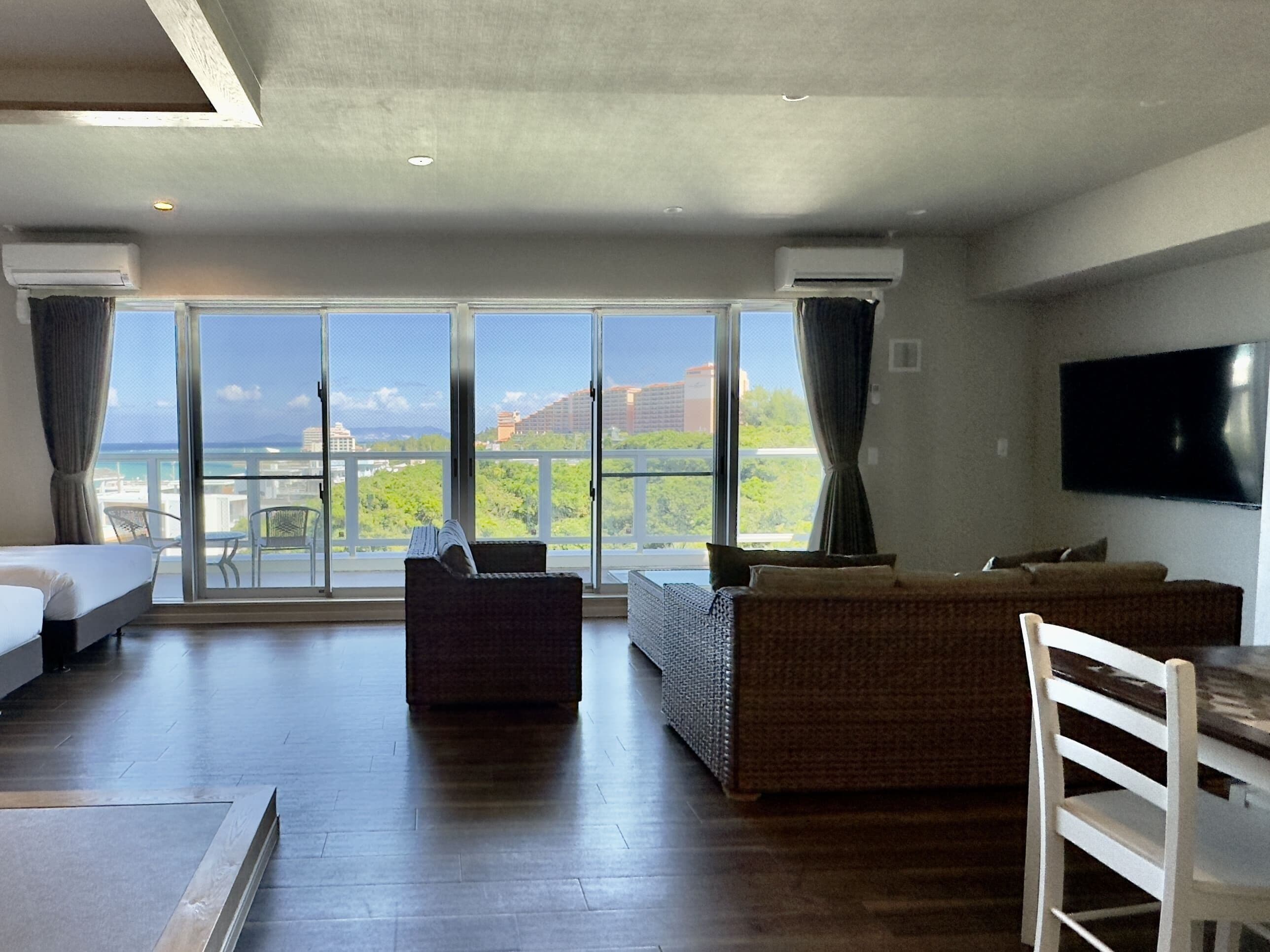 A spacious living area with a large window offering a view of the city and greenery. There is a sofa, a chair, and a flat-screen TV mounted on the wall.