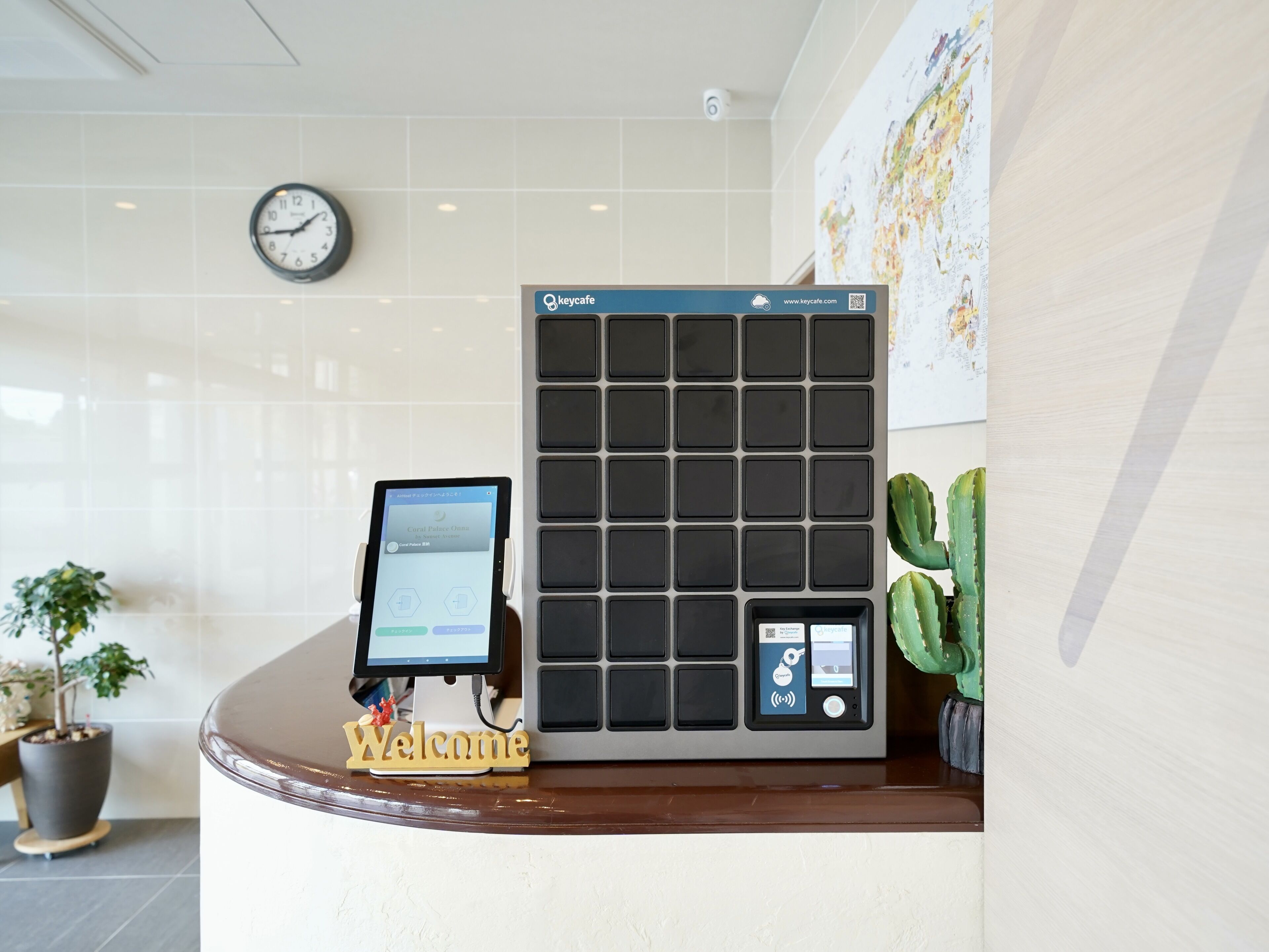 Un mostrador de recepción de hotel con un quiosco digital, un reloj, un cartel de bienvenida y una planta de cactus.