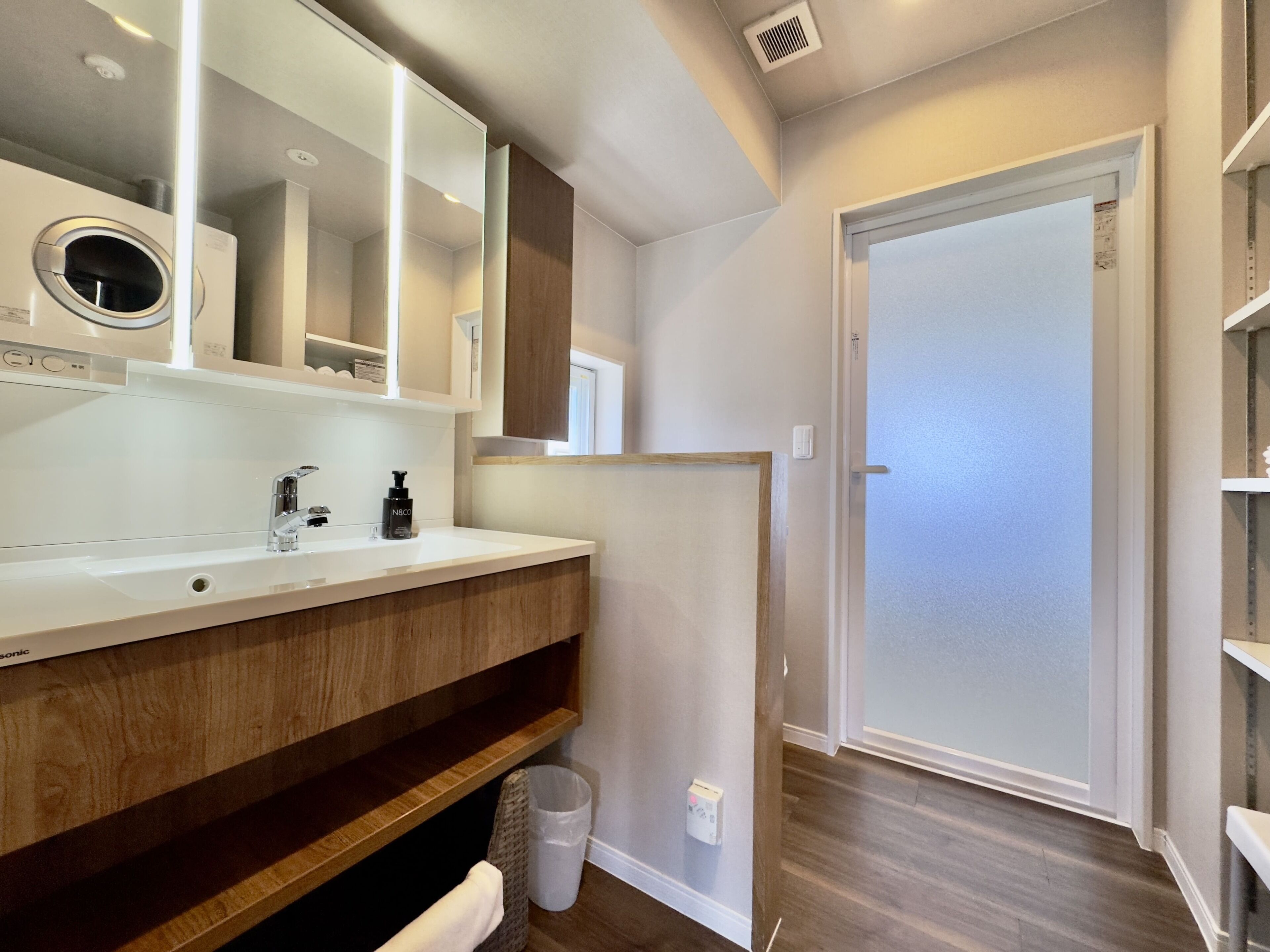 A modern bathroom with a white sink, mirror, and wooden vanity. There is a trash can and a door leading to another room.