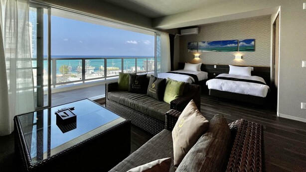 A hotel room with a balcony, a sofa, a bed, and a glass coffee table. - Coral Palace Onna by Sunset Avenue (Onna)