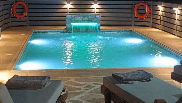 A rectangular indoor pool with a waterfall feature, surrounded by lounge chairs and towels. - Villa Parisi Naxos Deluxe 2 (Ναξος Αγκίδια)