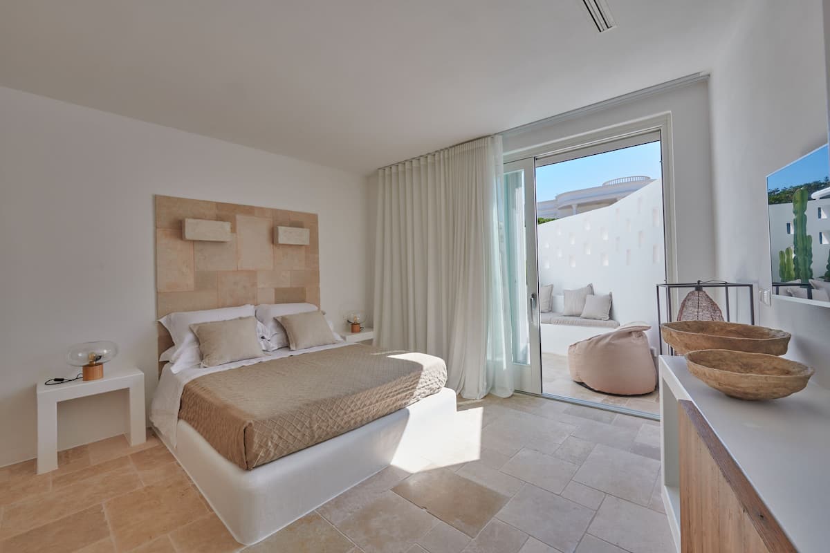 A modern bedroom with a large bed, a bedside table, a nightstand, and a view of the outside through a sliding glass door.