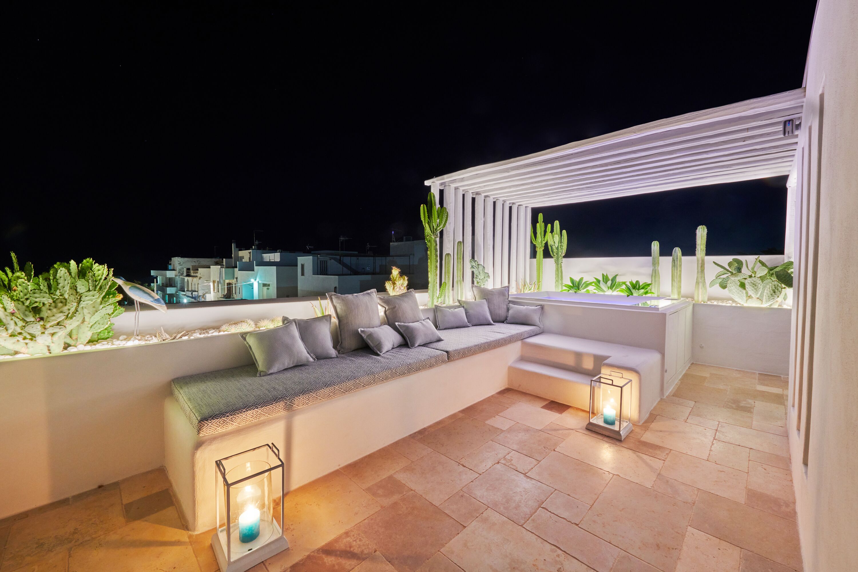 Une terrasse sur le toit avec un espace salon doté de coussins blancs, une pergola et des plantes en pot.