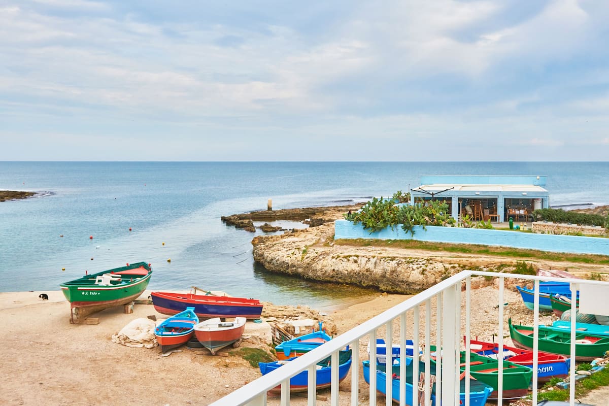 A coastal scene with boats docked on a sandy beach, a rocky outcrop, and a building with a blue roof.