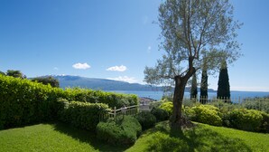 A well-maintained garden with a wooden fence, a tree, and a view of a lake and mountains in the distance. - Residence Limoni del Rustichel: Apartment N.1 with lake view, underground parking and pool. (Toscolano Maderno)