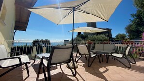 An outdoor patio with a table and chairs, a large umbrella, and a view of the sea. - Residence Limoni del Rustichel: Apartment N.1 with lake view, underground parking and pool. (Toscolano Maderno)