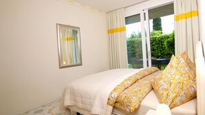A bedroom with a bed, a window with curtains, a small table with a lamp, and a view of greenery outside. - Residence Limoni del Rustichel: Apartment N.1 with lake view, underground parking and pool. (Toscolano Maderno)