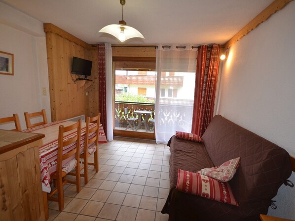 A cozy living room with a wooden dining table, chairs, a brown sofa with patterned cushions, and a TV mounted on the wall. - Charming Studio Near Village Center, 150m from Slopes, Balcony, Pet-Friendly (Les Gets)