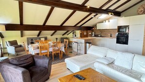 A modern loft apartment with exposed wooden beams, a dining area with wooden chairs, a white sofa, and a kitchen with white cabinets.
