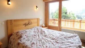 A bedroom with a wooden headboard, a quilted bedspread, two wall-mounted lights, and a large window with a view of greenery. - Charming Apartment Near Slopes with Balcony, Parking, and Modern Amenities (Les Gets)