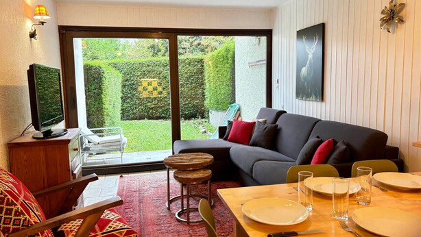 A cozy living room with a sofa, a dining table set for two, a television, and a view of a well-maintained garden through a sliding glass door. - Central Apartment in Les Gets, Steps from Village and Slopes (Les Gets)
