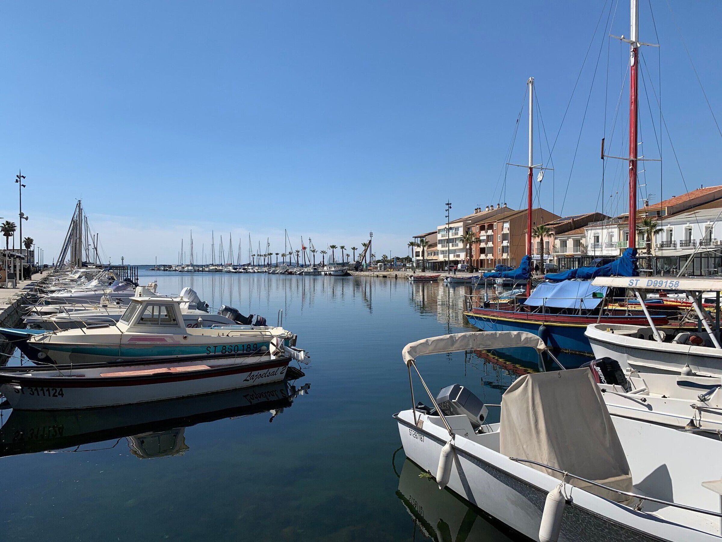 Un puerto deportivo con varias embarcaciones amarradas, cielo despejado y edificios al fondo.