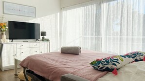Un dormitorio con una cama, un televisor sobre una cómoda y una ventana con cortinas de gasa.