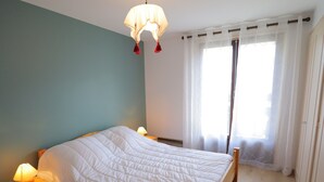 Ein Schlafzimmer mit einem Bett, Nachttischlampen, einem Holennachttisch, einem Fenster mit transparenten Vorhängen und einer Pendelleuchte.