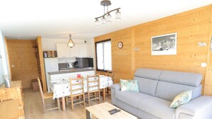 Ein gemütliches Wohnzimmer mit grauem Sofa, einem Holzesstisch und Stühlen. Im Hintergrund ist eine Küche mit weißen Schränken und einer Holzwand zu sehen.