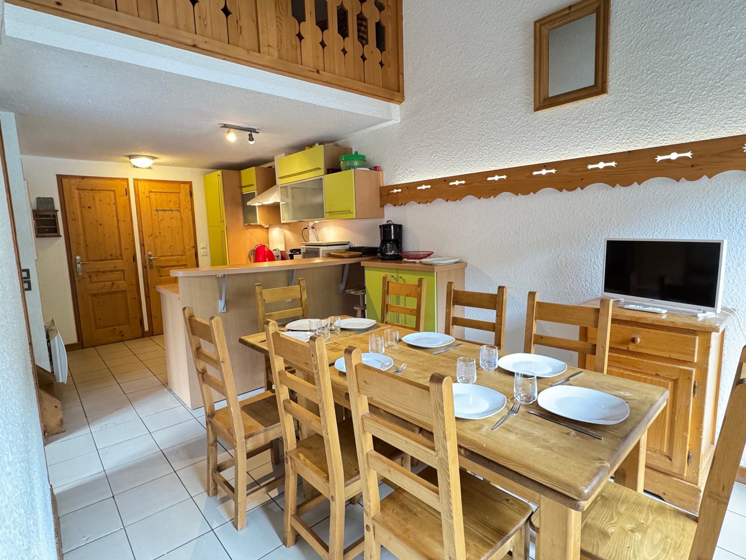 A dining area with a wooden table and chairs, a kitchen in the background, and a television on a wooden stand.