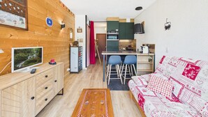 A cozy room with a wooden wall, a TV, a small cabinet, a bed with patterned bedding, and a kitchen area with a red curtain. - Ski-In/Ski-Out Studio with Pool, Tennis & Mountain Views - 1km from Les Gets Center (Les Gets)