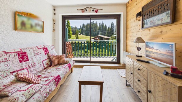 A cozy living room with a sofa, a table, and a TV on a wooden cabinet. - Ski-In/Ski-Out Studio with Pool, Tennis & Mountain Views - 1km from Les Gets Center (Les Gets)