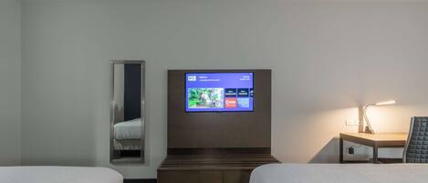 A hotel room with two beds, a TV mounted on the wall, a bedside table with a lamp, and a small cabinet.