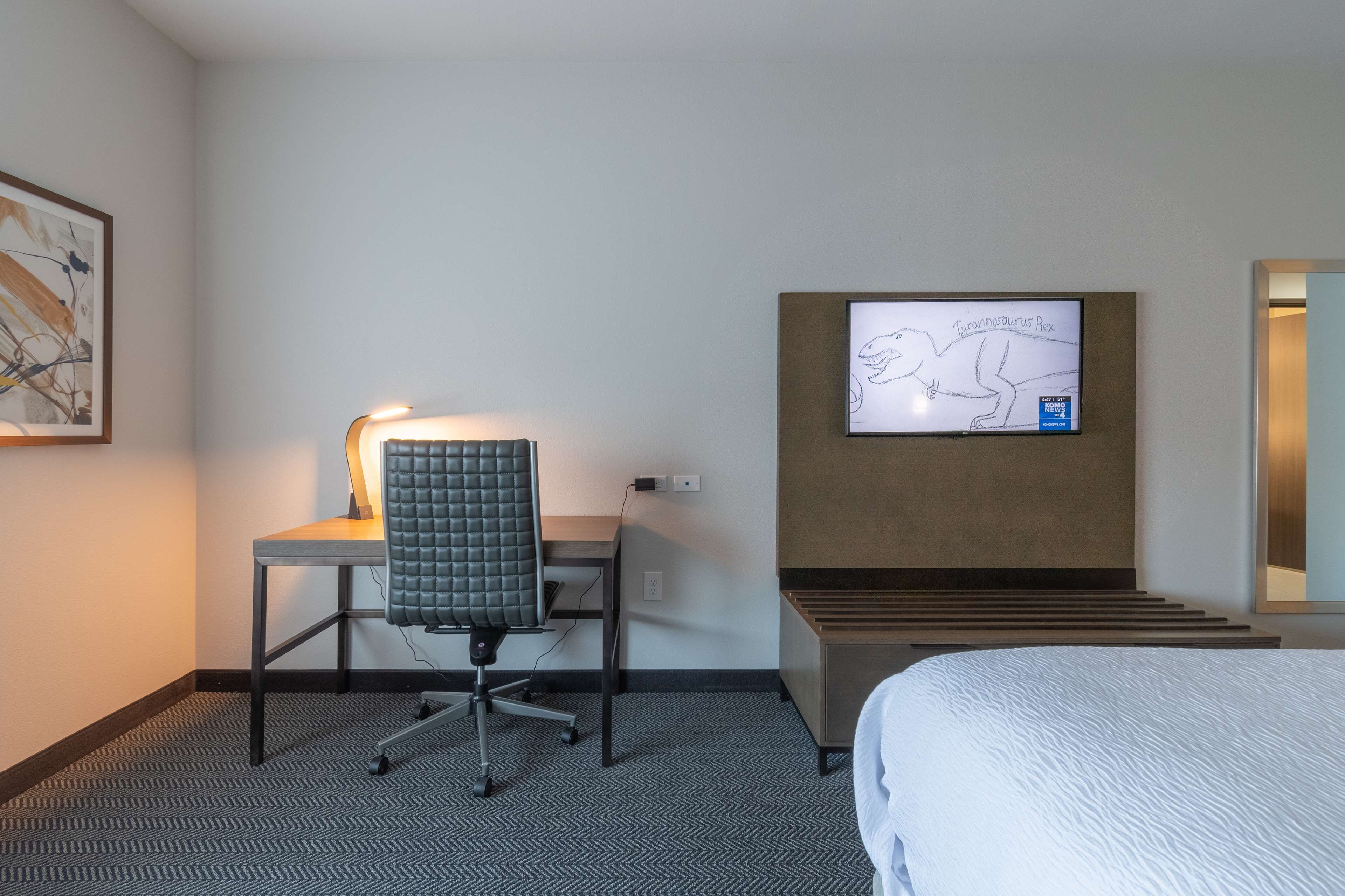 Una habitació d'hotel amb un llit, una taula de treball amb una cadira, una televisió penjada a la paret i un quadre amb marc a la paret.