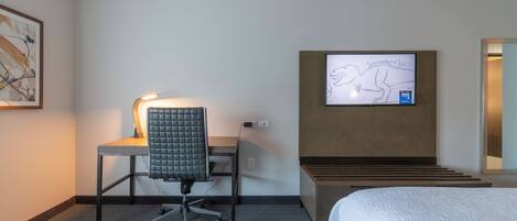 A hotel room with a bed, a desk with a chair, a television mounted on the wall, and a framed picture on the wall.
