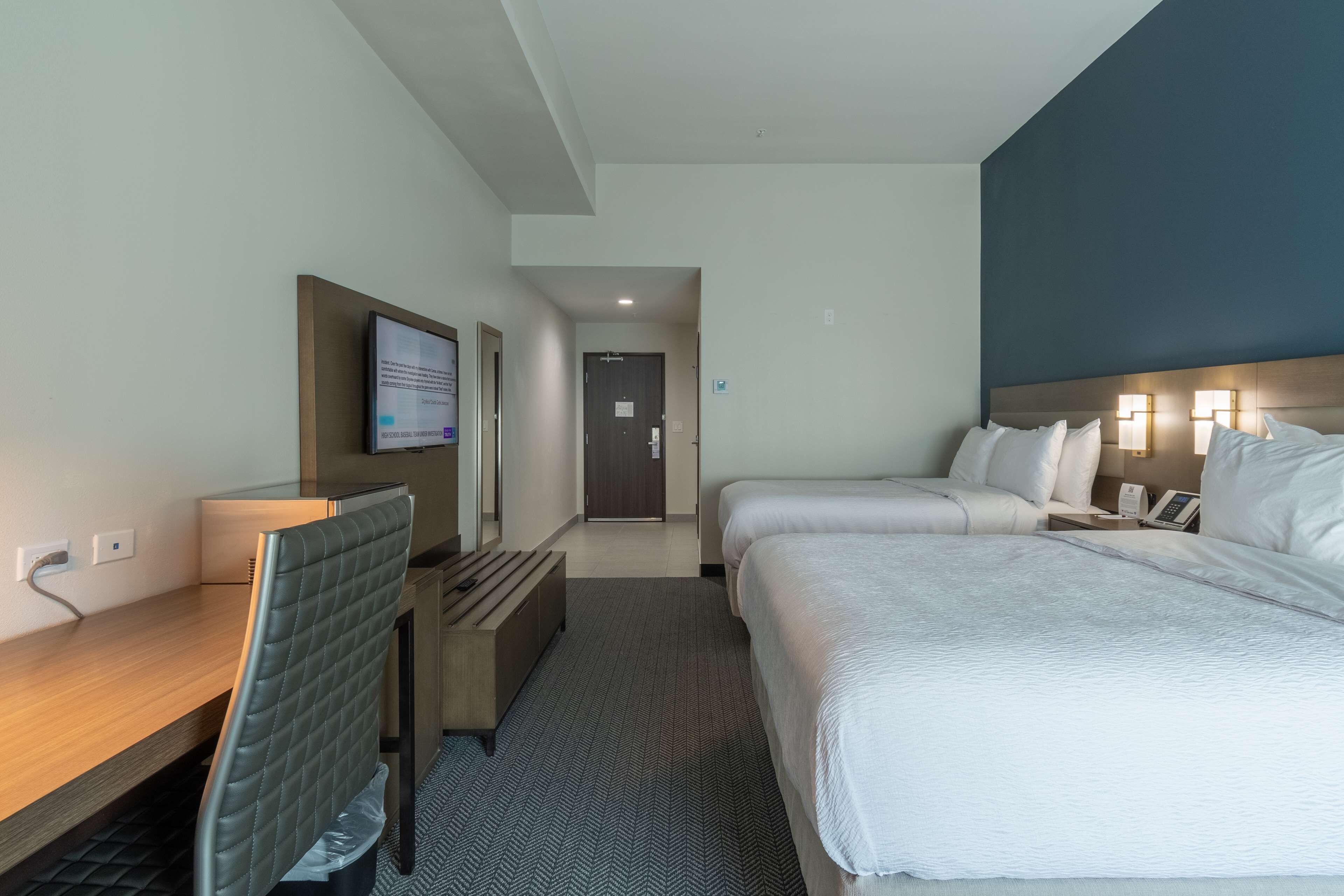 A hotel room with a desk, two beds, a TV, and a door leading to a hallway.